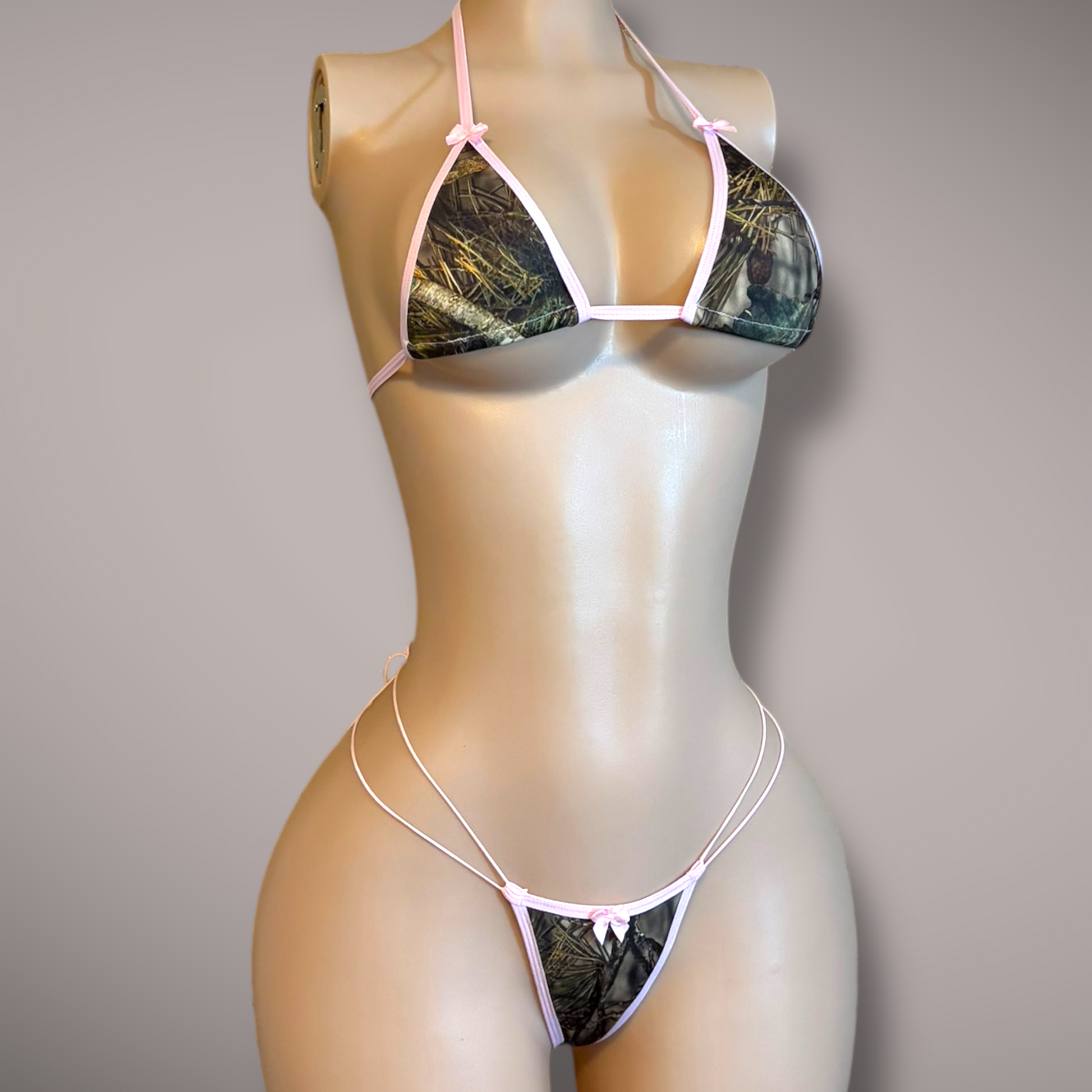 Angled front view of a camo and pink micro bikini on a mannequin, highlighting the triangle top, small bows, and double-strap thong bottom.